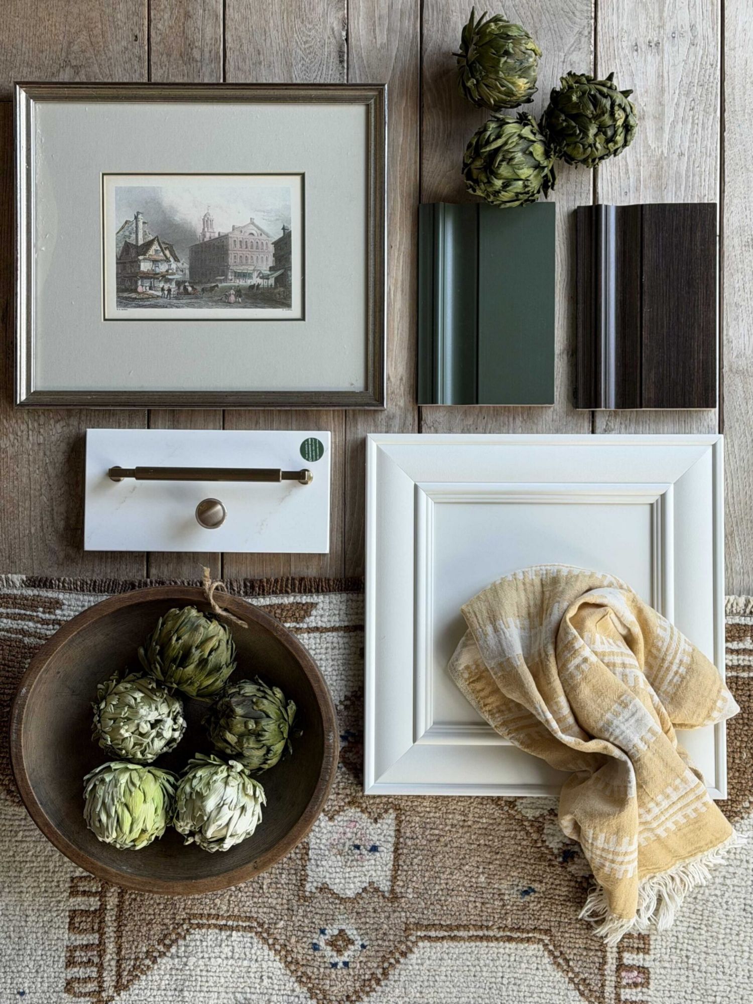 Cabinetry design flat lay: green and walnut door samples, a white painted cabinet door, brass hardware on a quartz sample, a framed print, and a bowl of dried artichokes